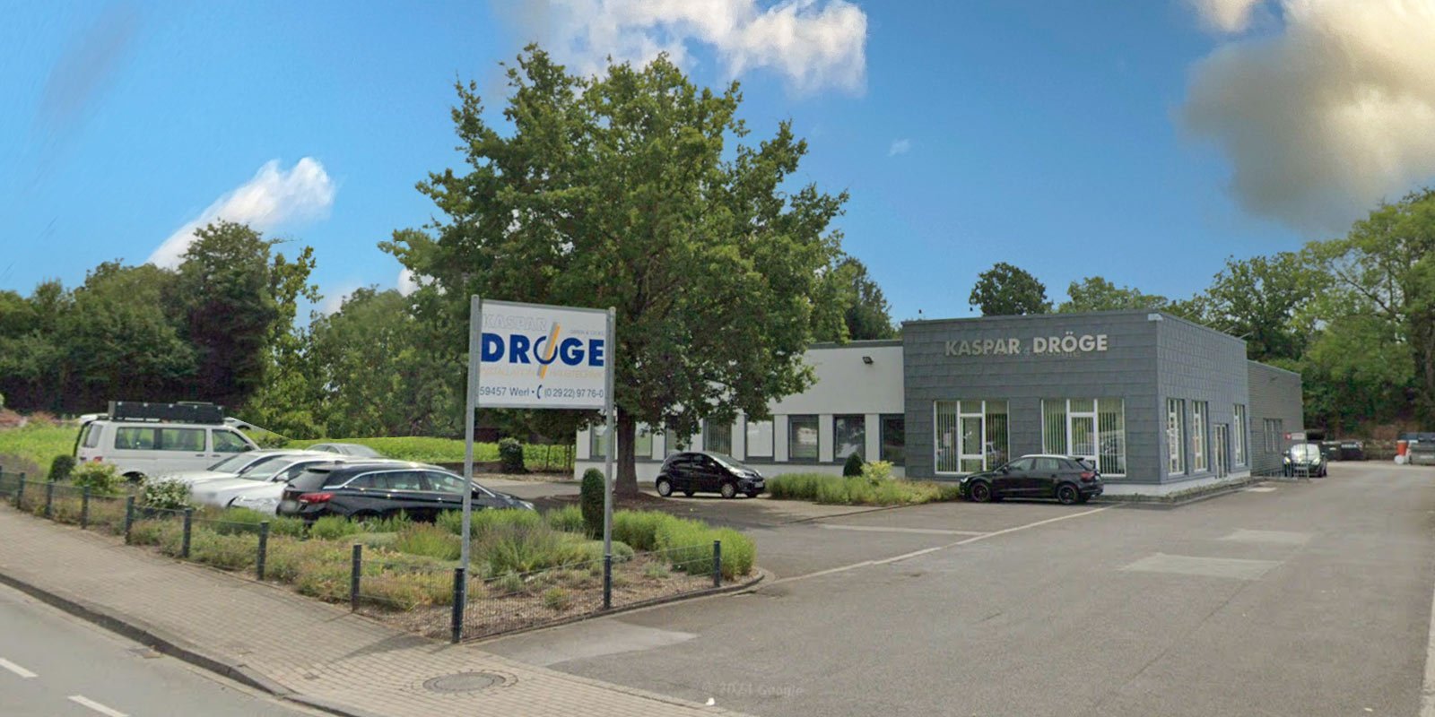 Das moderne Gebäude mit dem Schild 'KASPAR DRÖGE' steht in einer ruhigen Umgebung, umgeben von Bäumen und Autos.